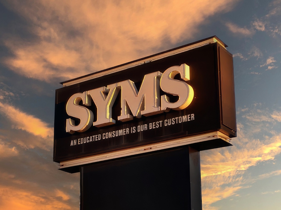 Photo of a sign saying "SYMS" in large letters, with "AN EDUCATED CONSUMER IS OUR BEST CUSTOMER" in smaller writing underneath